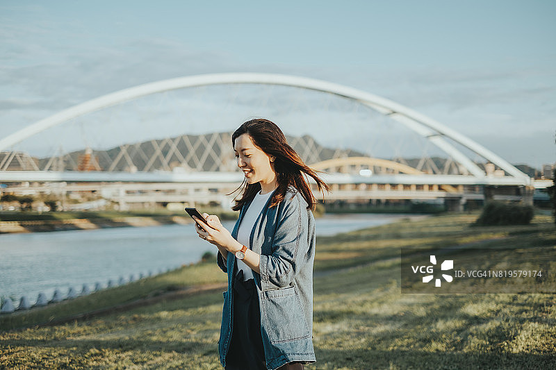 微笑的亚洲女士拿着智能手机在公园里放松，站在河对岸的桥上图片素材