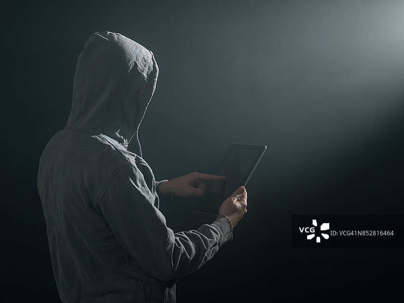 一名男子穿着连帽衬衫，在黑暗中使用数字平板电脑入侵图片素材