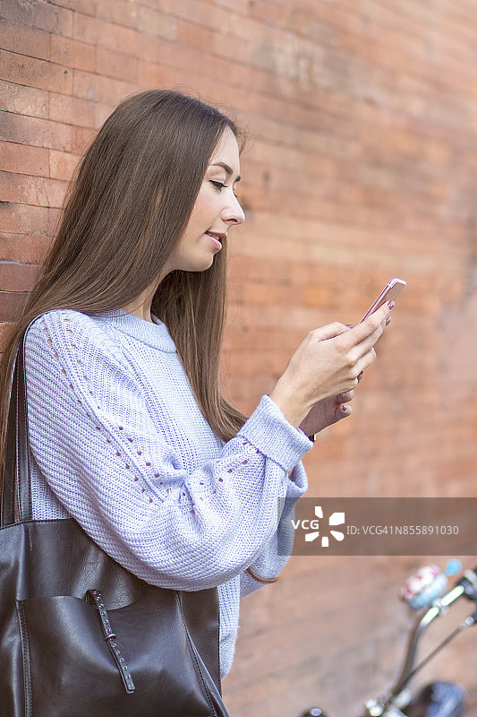 年轻女子靠在红砖墙上用手机。图片素材