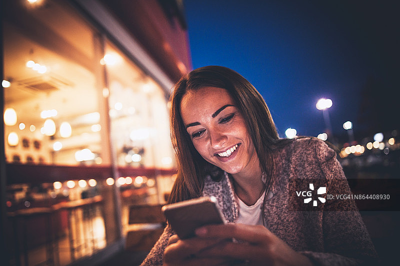 微笑的年轻女子使用智能手机在夜晚的街道上图片素材