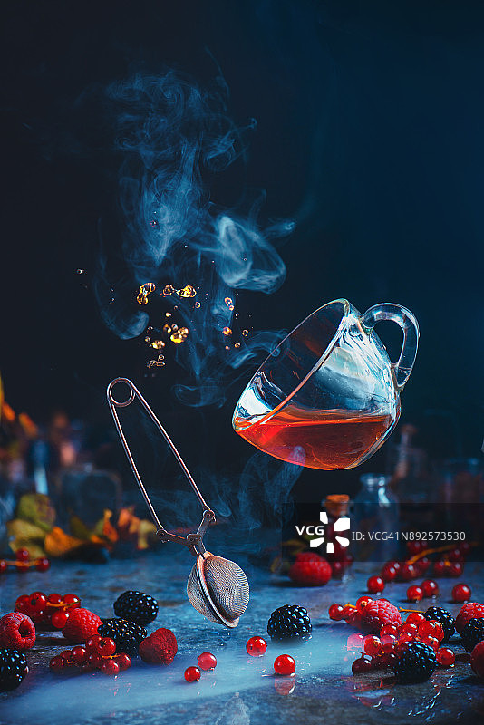 悬浮透明的茶杯与茶过滤器和飞行滴在一个黑色的背景与醋栗，覆盆子，黑莓和薄荷叶。图片素材