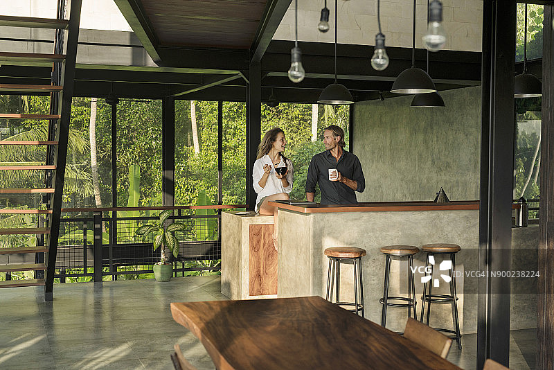在被茂密的热带花园包围的玻璃立面的现代设计厨房里，一对夫妇在一起吃早餐，互相微笑图片素材