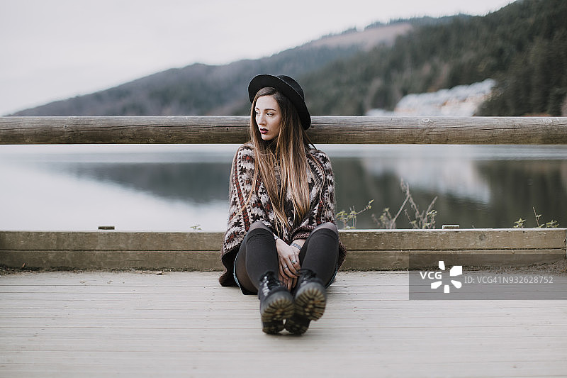 一幅时尚的年轻女子戴着帽子和斗篷坐在木桥上的肖像图片素材
