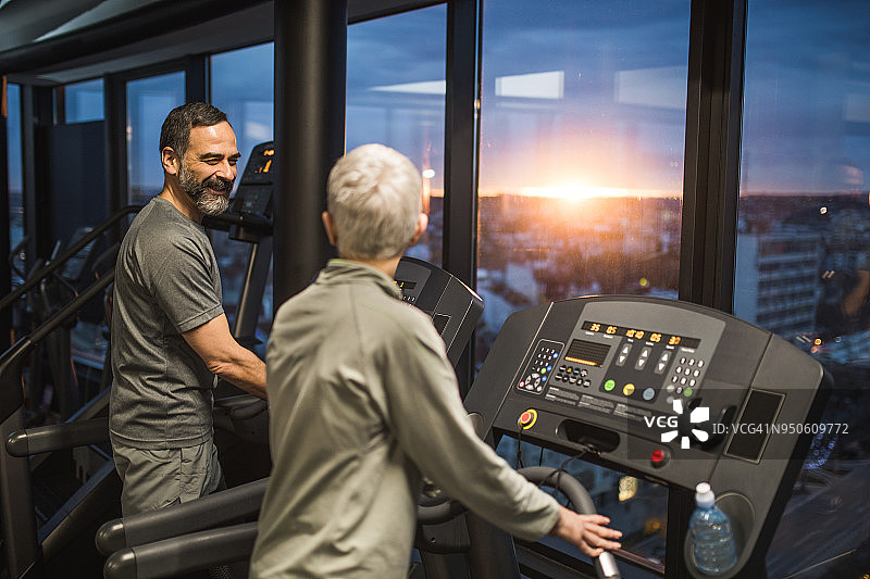 一个快乐的老人在健身房里跑步机上和一个女人聊天。图片素材