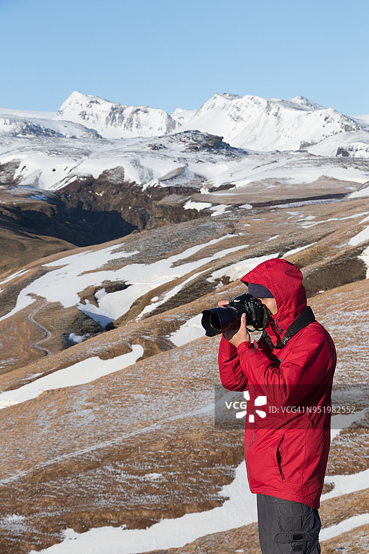 在前往冰岛南部维克市的路上，一名身穿红色夹克的男摄影师在视点拍摄雪山图片素材