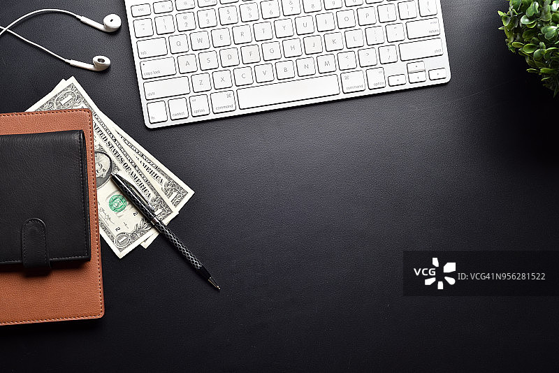 美元躺在黑色皮革桌子上，电脑键盘，笔记本，钱包，耳机，小绿色桌子植物拷贝空间图片素材