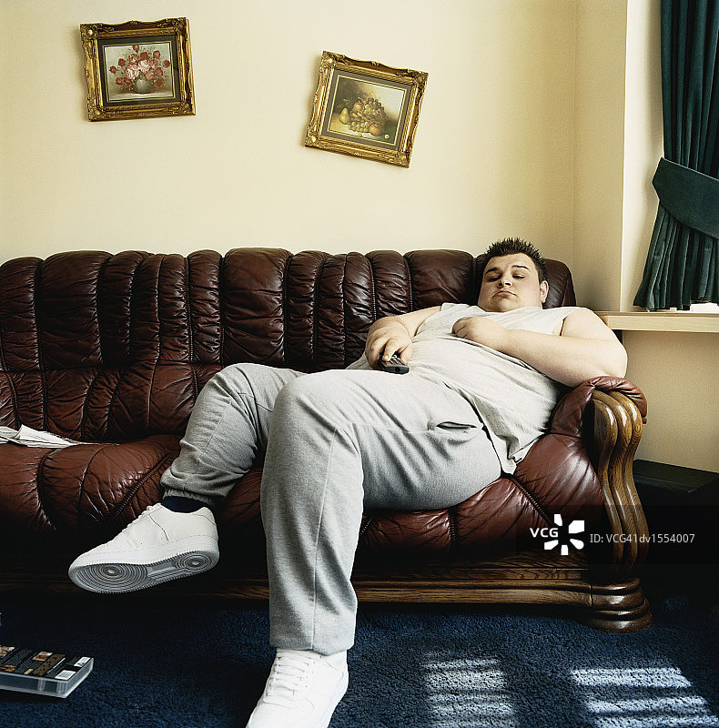 一个超重的年轻人躺在沙发上看电视睡着了图片素材