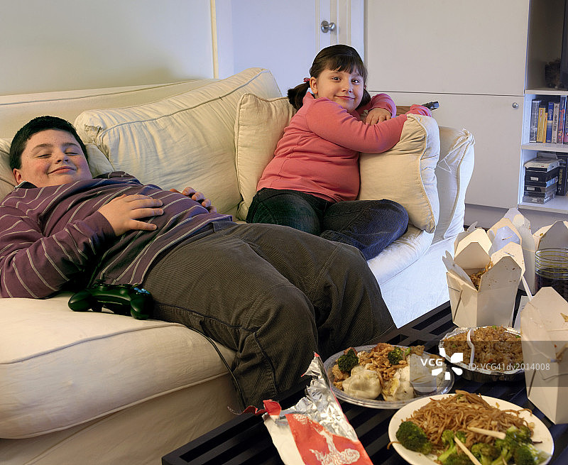 超重的哥哥和妹妹坐在沙发上吃外卖和看电视图片素材