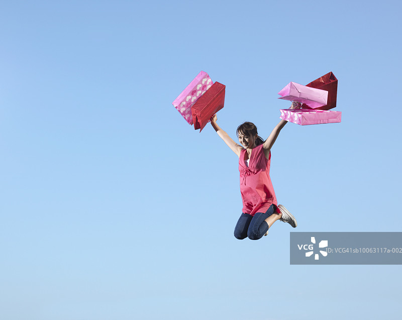 年轻女子拿着购物袋在蓝天上跳跃，微笑着图片素材