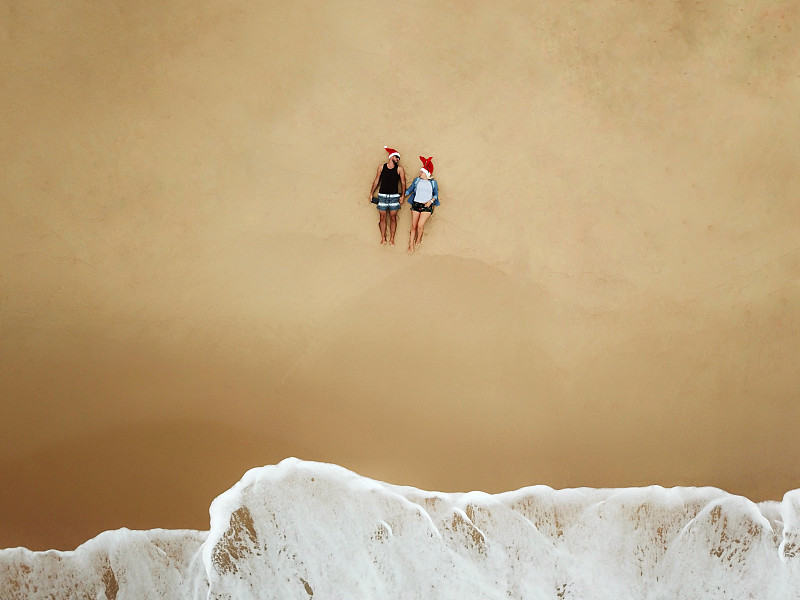 这是一对戴着圣诞老人帽坐在海滩上的年轻夫妇的航拍照片摄影图片