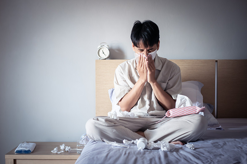 亚洲男人感冒了，坐在舒适的床上用纸巾擦鼻涕。生病在家图片下载