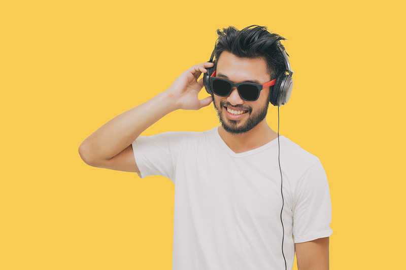 亚洲英俊的男人，有胡子，微笑和大笑，用智能手机听音乐的黄色背景耳机图片素材