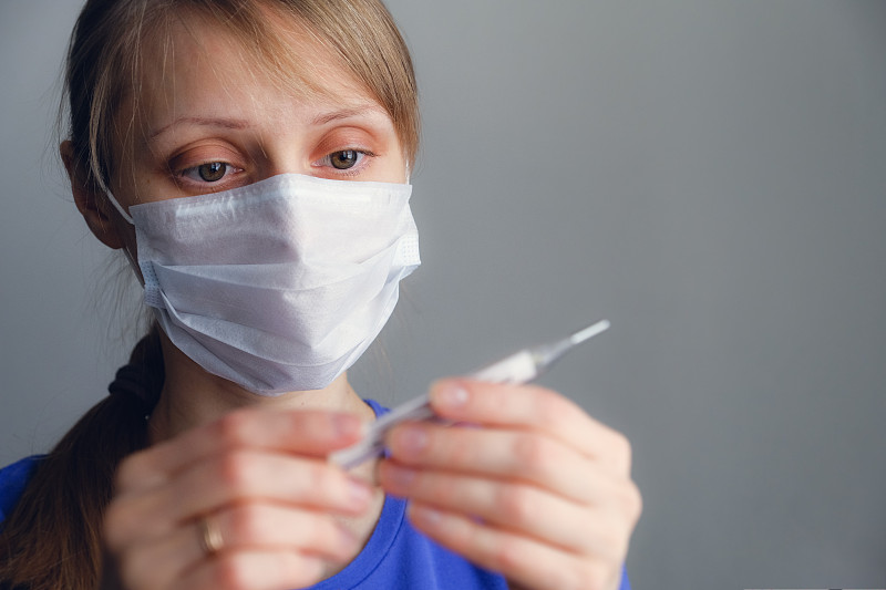 健康的概念。一个生病的女孩戴着一次性医用口罩看温度计。关闭。女孩检查了体温。流感季节，流感病毒。削弱了免疫力。图片下载