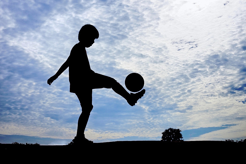 日落时分，在多云的天空下，一个男孩在球场上踢足球图片素材