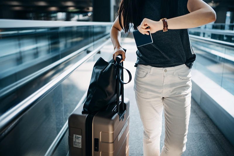 年轻的亚洲女子拿着行李箱和护照走在机场大厅时，手表上显示时间图片下载
