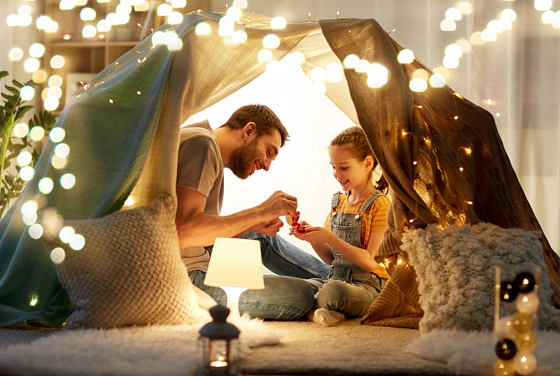 家庭、hygge和人们的概念——快乐的父亲和小女儿晚上在家里的孩子帐篷里玩茶会。家庭在孩子们的帐篷里玩茶话会图片下载