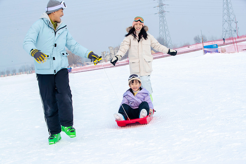 滑雪场上父母和坐在雪上滑板的女儿玩耍图片素材