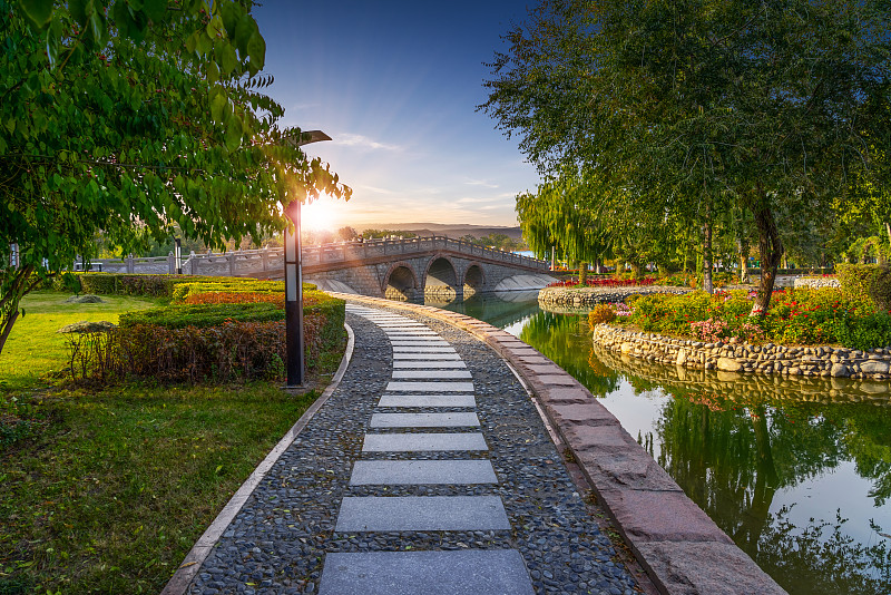 新疆乌鲁木齐人民公园湖景石材拱桥曲线步道日落逆光图片素材