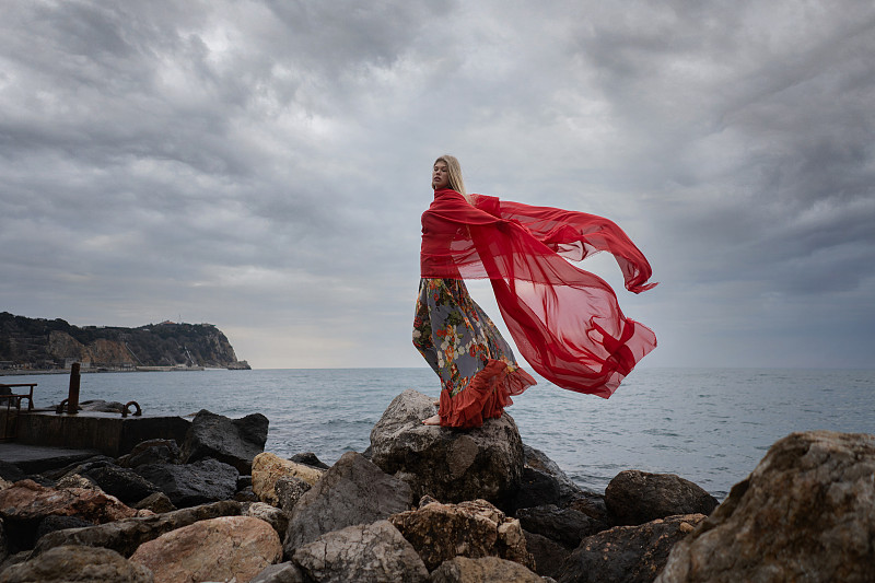 一个穿红衣服的女人站在海边的岩石上，对着天空图片下载