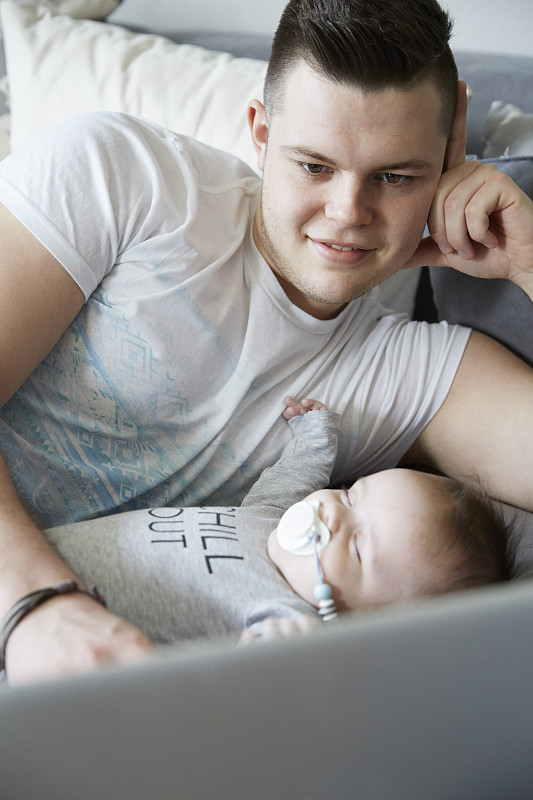 年轻的父亲使用笔记本电脑，婴儿睡在沙发上图片下载