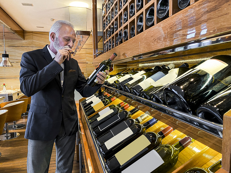 经验丰富的年长侍酒师选择一个葡萄酒品尝在一个豪华的酒窖。他在抽雪茄图片下载