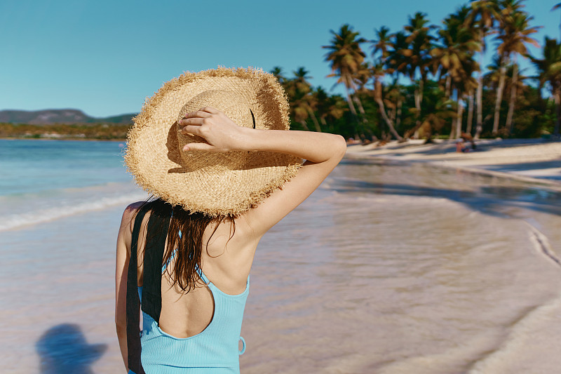 在一个热带岛屿上，一名身穿泳衣的年轻女子手戴一顶草帽，沿着沙滩散步，背景是棕榈树图片下载