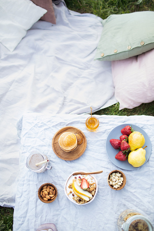 夏季健康素食野餐在公园。
水果，可可，蜂蜜煎饼和有机格兰诺拉麦片在亚麻桌布上装饰枕头和柳条篮子。图片素材