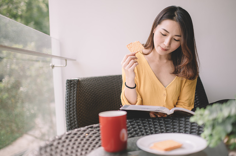 一位亚洲华裔美女中午在她的阳台上看书，放松，享受她的数字排毒时间，喝着咖啡，吃着饼干当点心图片素材