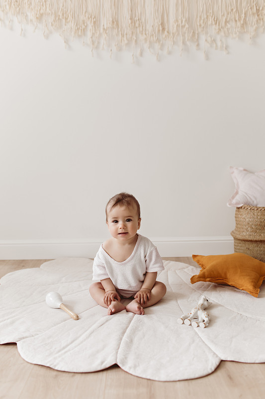 可爱的微笑蹒跚学步坐在贝壳地毯上。
可爱的婴儿与生态玩具，星星枕头和棉流苏坐在地板上美丽温柔的游戏室。图片下载