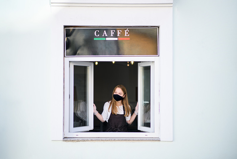 戴着面罩和手套的年轻侍者通过咖啡店窗口端咖啡。图片下载