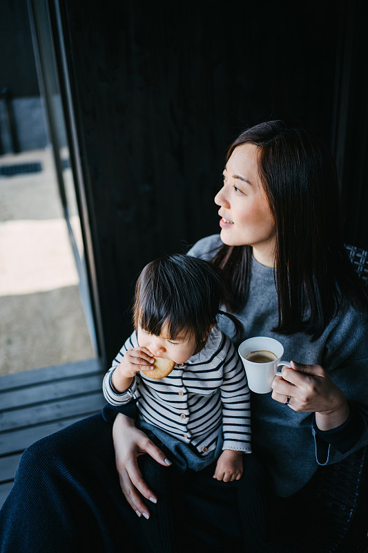 可爱的亚洲小女孩坐在妈妈的腿上吃饼干，妈妈喝咖啡，他们在阳台上放松，享受亲密的家庭时光图片下载