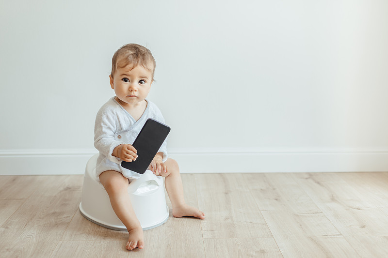 有趣的幼儿坐在白色便盆椅上玩手机。图片素材