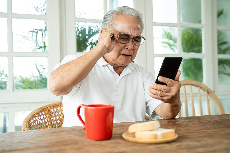 年长的亚洲男性有视力问题，在家里吃早餐时戴着眼镜盯着智能手机看信息。在室内视力模糊的老人图片素材