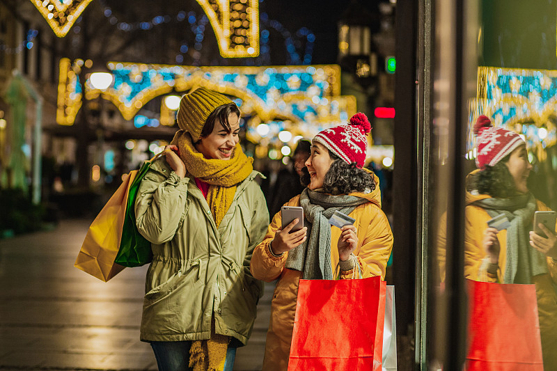 Two excited female friends in a Christmas shopping using credit card for mobile paying图片素材