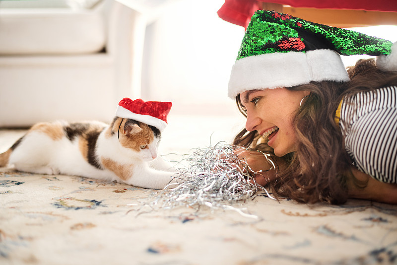 年轻女子和她的猫戴着圣诞老人的帽子在家里一起玩图片下载