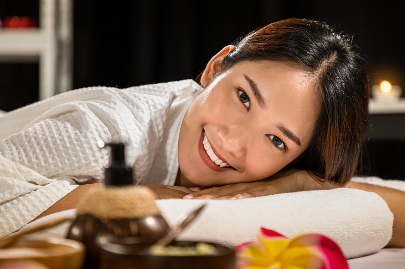 美丽年轻的亚洲女人休息在床上水疗，与按摩油，磨砂粉，和蜡烛，看着相机。图片下载