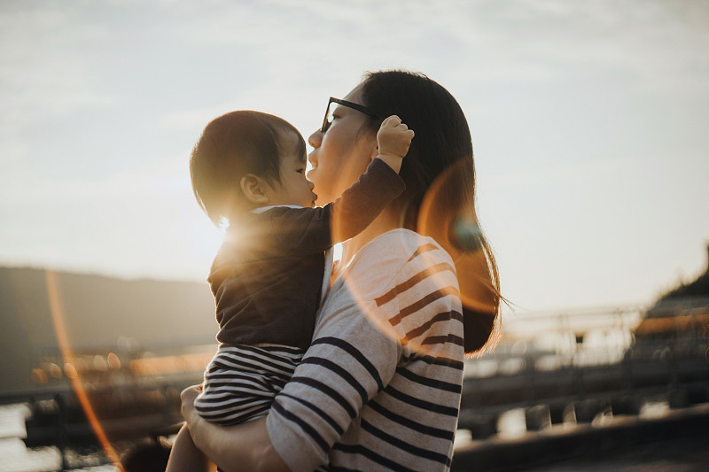 一位慈爱的年轻亚洲母亲抱着可爱的小女儿，在日落时分在公园里悠闲地散步。享受时刻和高质量的家庭团聚时间在一起。亲情与生活方式图片下载