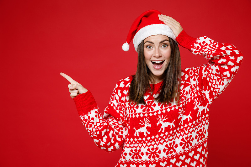 兴奋的年轻圣诞女人20多岁，穿着毛衣，戴着圣诞帽，食指指向一边，在模拟拷贝空间，手放在头上孤立在红色背景上。新年庆祝快乐节日概念。图片下载