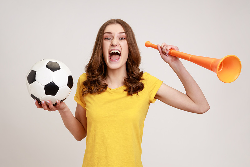 这是一个年轻快乐的女人的肖像，棕色头发，穿着黄色的t恤，拿着大扩音器和足球，庆祝冠军的开始。图片下载