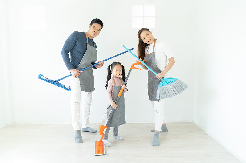 年轻快乐的亚洲家庭在客厅里打扫房间，玩拖把和扫帚。带着乐趣，看着镜头。人，爱，家庭，家政和家庭清洁的理念。图片下载