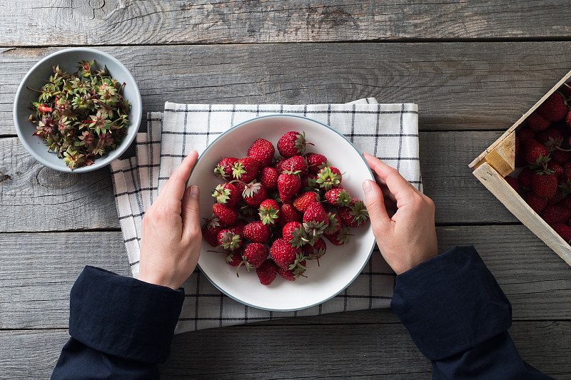 成熟的红色草莓放在一个碗或陶瓷盘子里，放在木制背景或桌子上。一个女人或一个女孩手里有一个大浆果，她会吃掉它。素食的概念，纯素和生食的营养和饮食。从农场到餐桌。蔬菜食品。摄影图片
