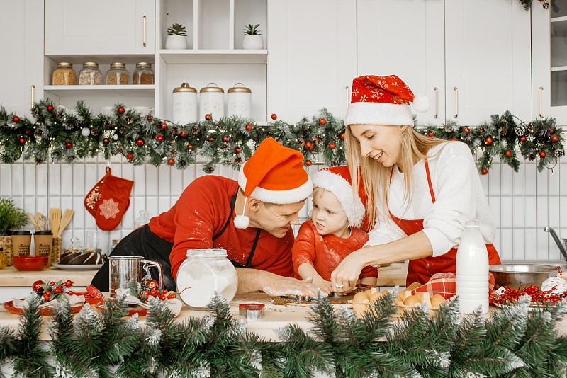 幸福快乐的一家人戴着圣诞老人的帽子，用厨房里的饼干切刀把生面团切成饼干。自制蛋糕，圣诞糖果，寒假概念。图片下载