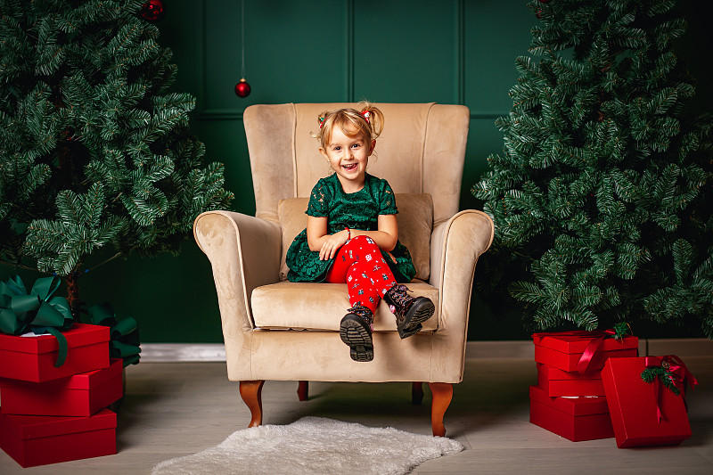 可爱的金发小女孩坐在大扶手椅之间的圣诞树和红色的礼品盒图片下载