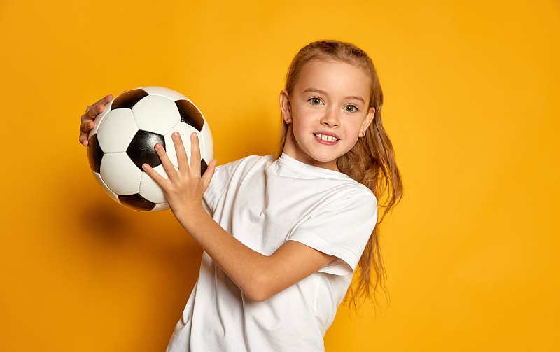 小美丽的女孩的肖像，孩子在白色t恤摆姿势与足球孤立在黄色的工作室背景。成功图片下载