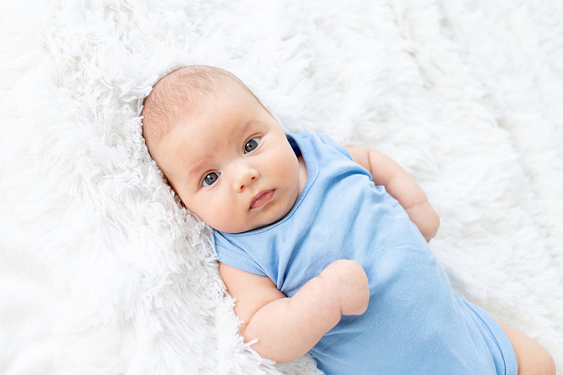 三个月大的可爱男婴，穿着蓝色紧身衣，躺在家里的白色床上图片下载