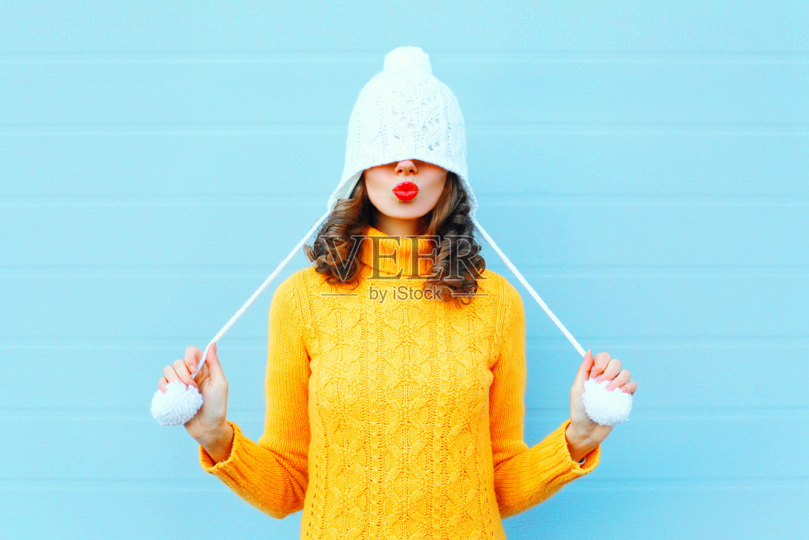 快乐酷女孩吹红嘴唇，让空气亲吻戴着针织帽，黄色毛衣在蓝色的背景照片摄影图片