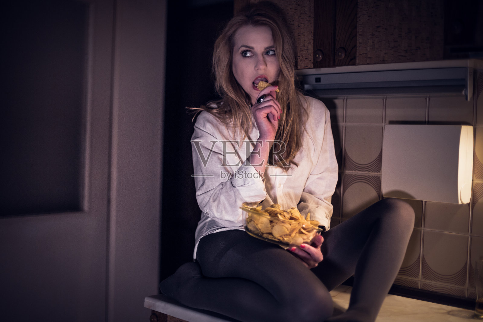 性感的年轻女人穿着睡衣坐在厨房的柜台上，在晚上吃土豆片照片摄影图片