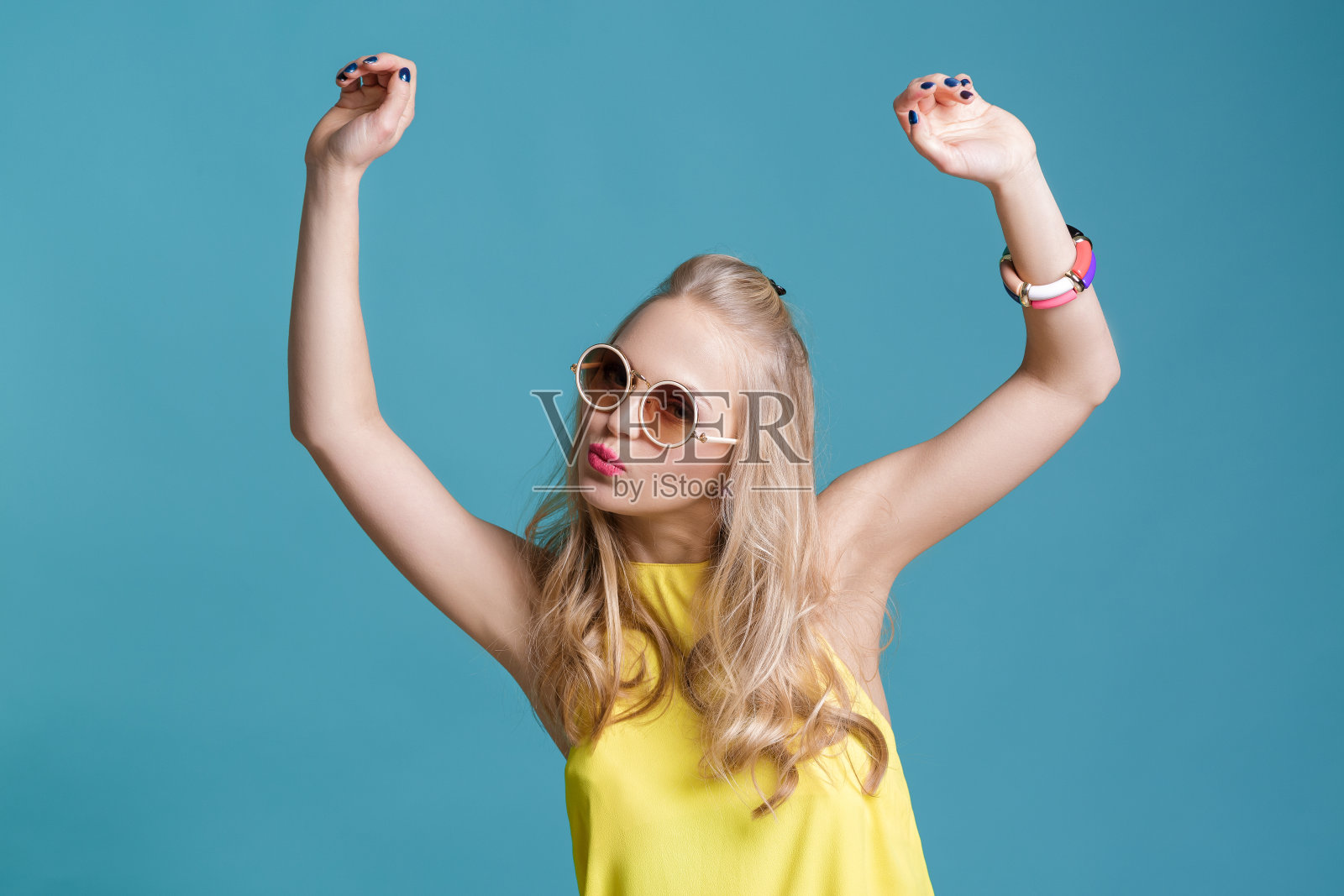 美丽的金发女人的肖像戴着太阳镜和黄色的衬衫在蓝色的背景跳舞。无忧无虑的夏天。照片摄影图片