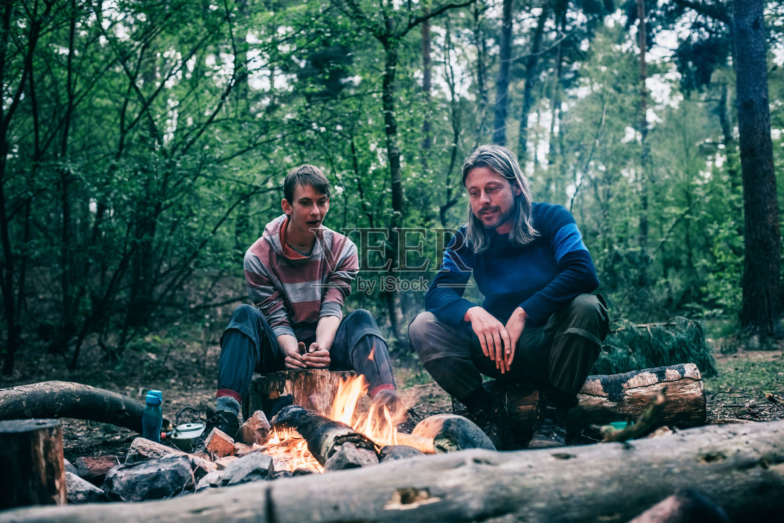 在春天的森林里，两个人围坐在篝火旁。照片摄影图片