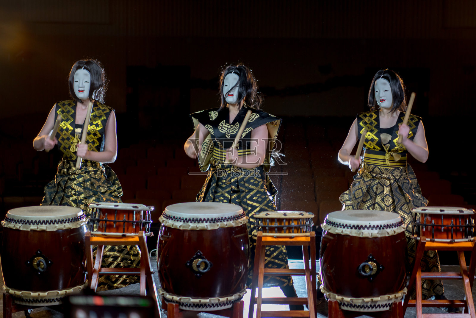 三个鼓手女孩戴着白色的恶魔面具和鼓槌，在黑暗的背景下拍摄。照片摄影图片